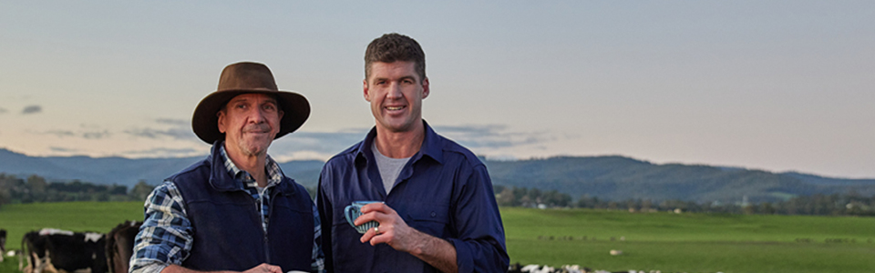 Jonathan Brown and farmer holding glasses of milk. Standing in paddock, cows behind them.